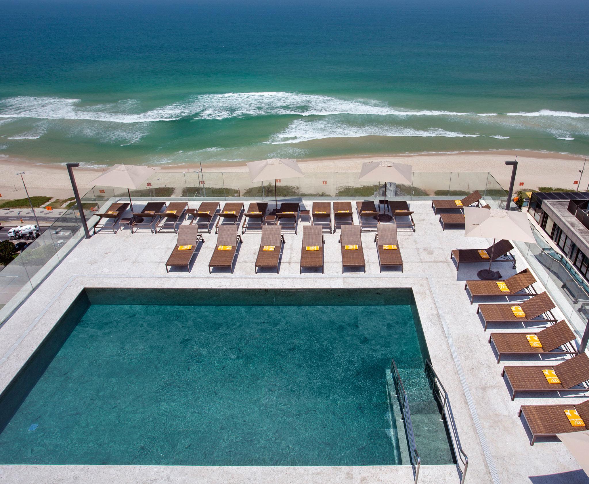 Windsor Marapendi Hotel Rio de Janeiro Exteriér fotografie The photo shows a rooftop area with a swimming pool and several lounge chairs arranged around it. The pool has a clear blue surface, and the lounge chairs are positioned in a neat row, some with yellow cushions. In the background, you can see a beaut