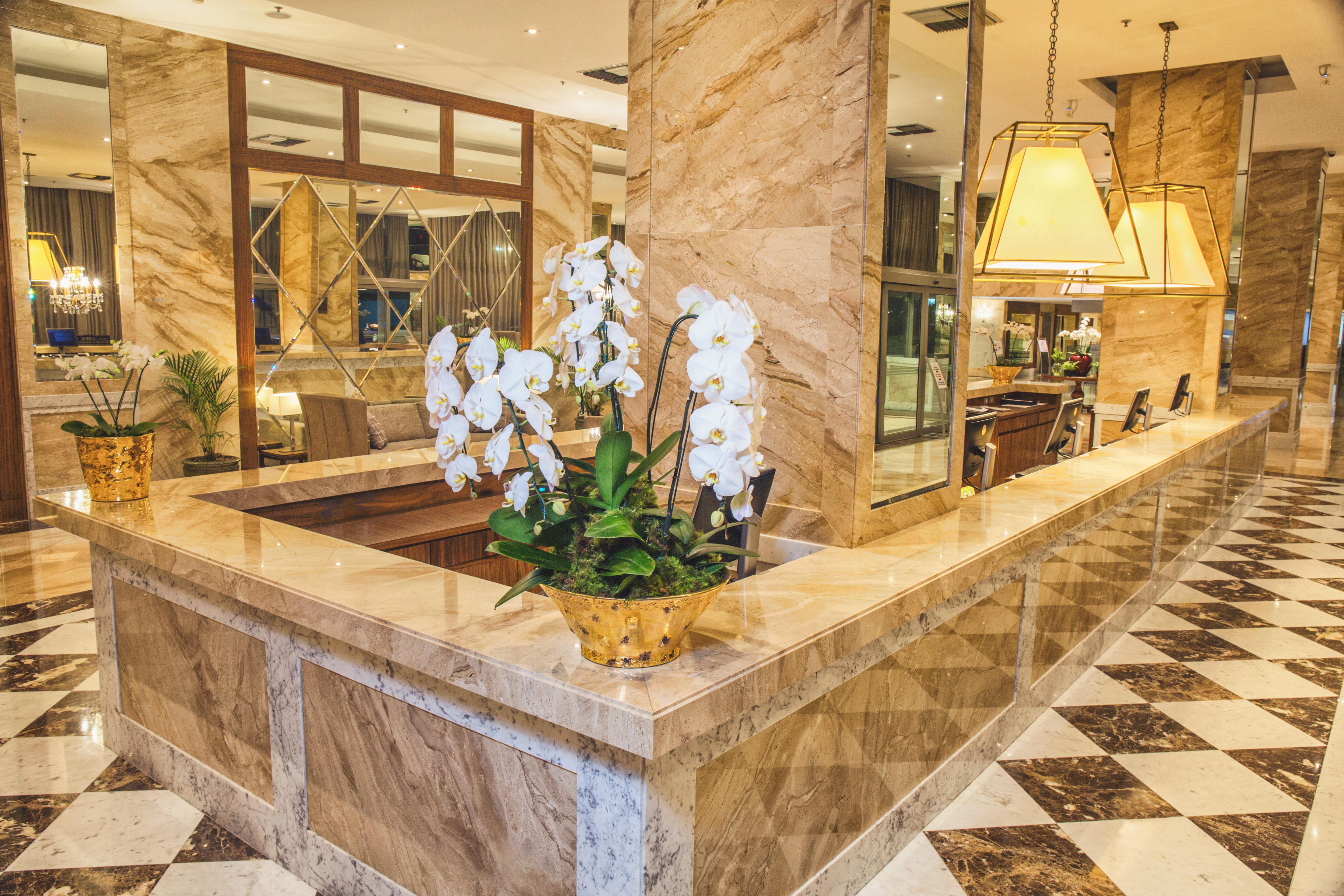 Windsor Marapendi Hotel Rio de Janeiro Exteriér fotografie The photo depicts a luxurious hotel lobby featuring a polished marble reception desk. There are elegant white orchids arranged in a gold pot on the counter, adding a touch of sophistication. The surrounding interior showcases marble walls, stylish li