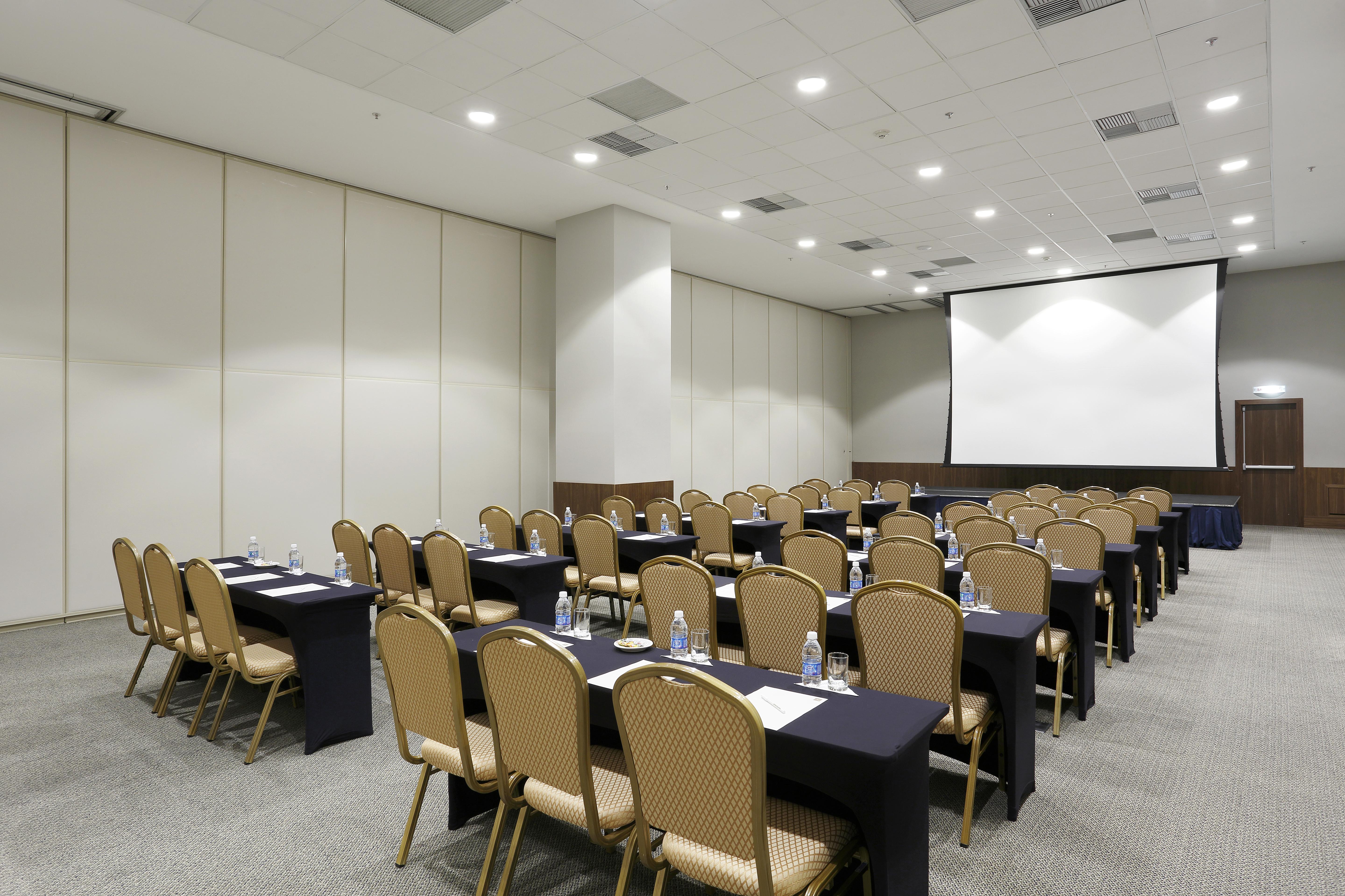 Windsor Marapendi Hotel Rio de Janeiro Exteriér fotografie The photo shows a conference or meeting room arranged with several rows of tables and chairs. The tables are covered with dark tablecloths and have water bottles placed on them. The room is bright and spacious, featuring a large projection screen on 