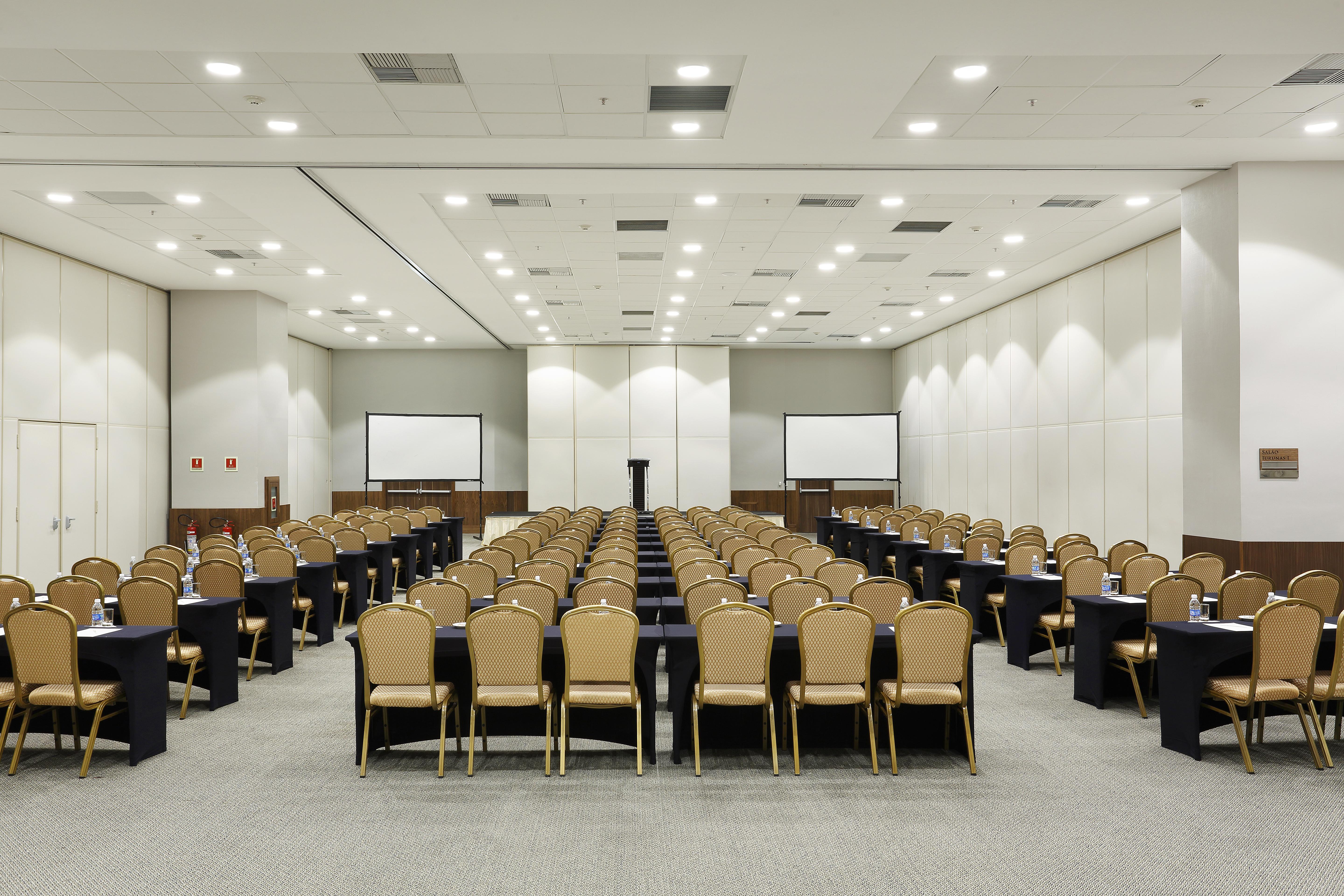 Windsor Marapendi Hotel Rio de Janeiro Exteriér fotografie The photo shows a conference room arranged for an event or meeting. There are rows of neatly arranged chairs with golden frames, paired with black tables. The room has bright overhead lighting and features two blank screens in the front, likely for p