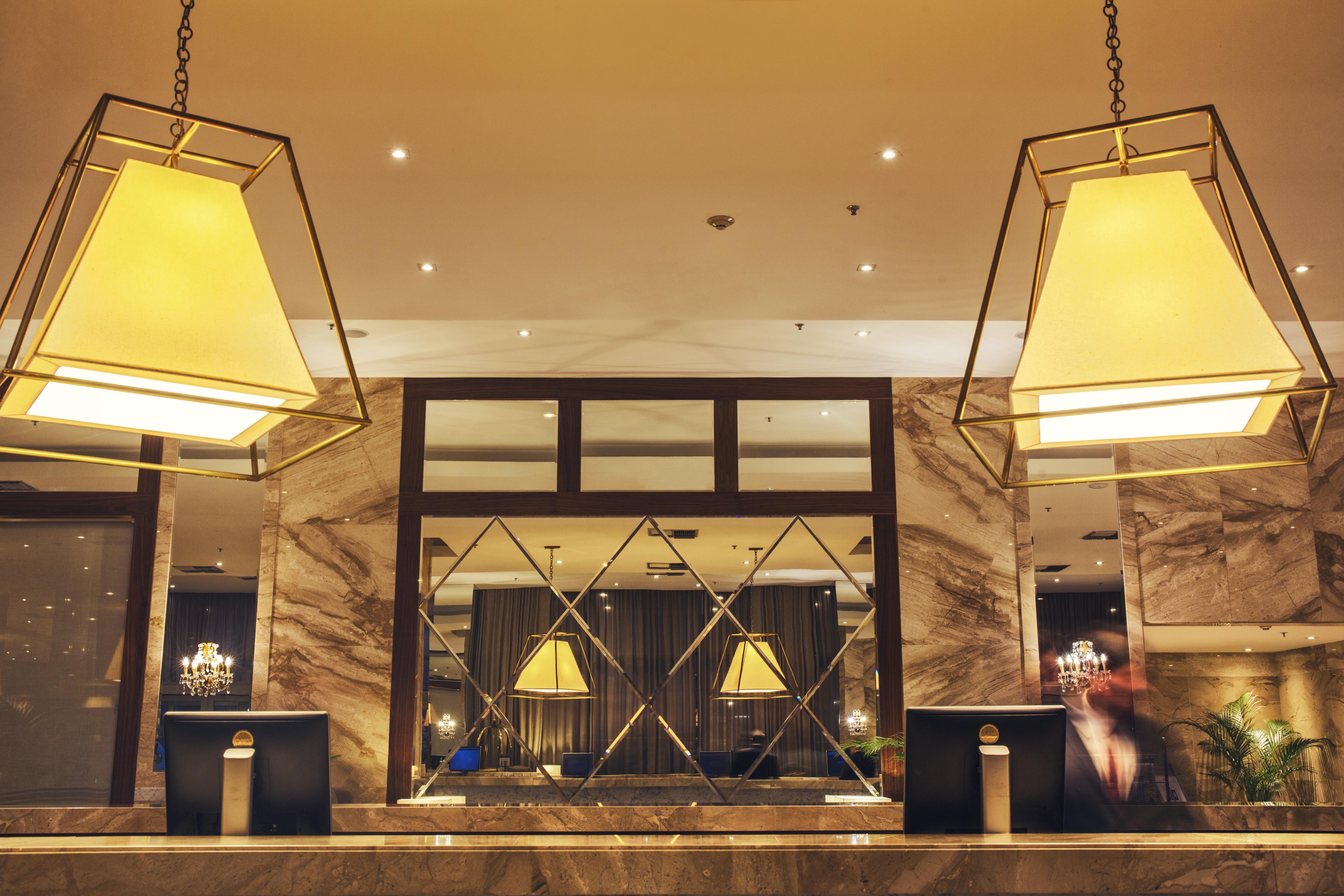 Windsor Marapendi Hotel Rio de Janeiro Exteriér fotografie The photo shows a hotel lobby or reception area. There are large geometric light fixtures hanging from the ceiling, providing a warm glow to the space. The reception desk is made of a polished material, likely marble, with decorative elements and two